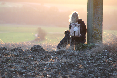 Frau mit Hund sitzt auf einem Wegestein und blickt auf dunstige Landschaft